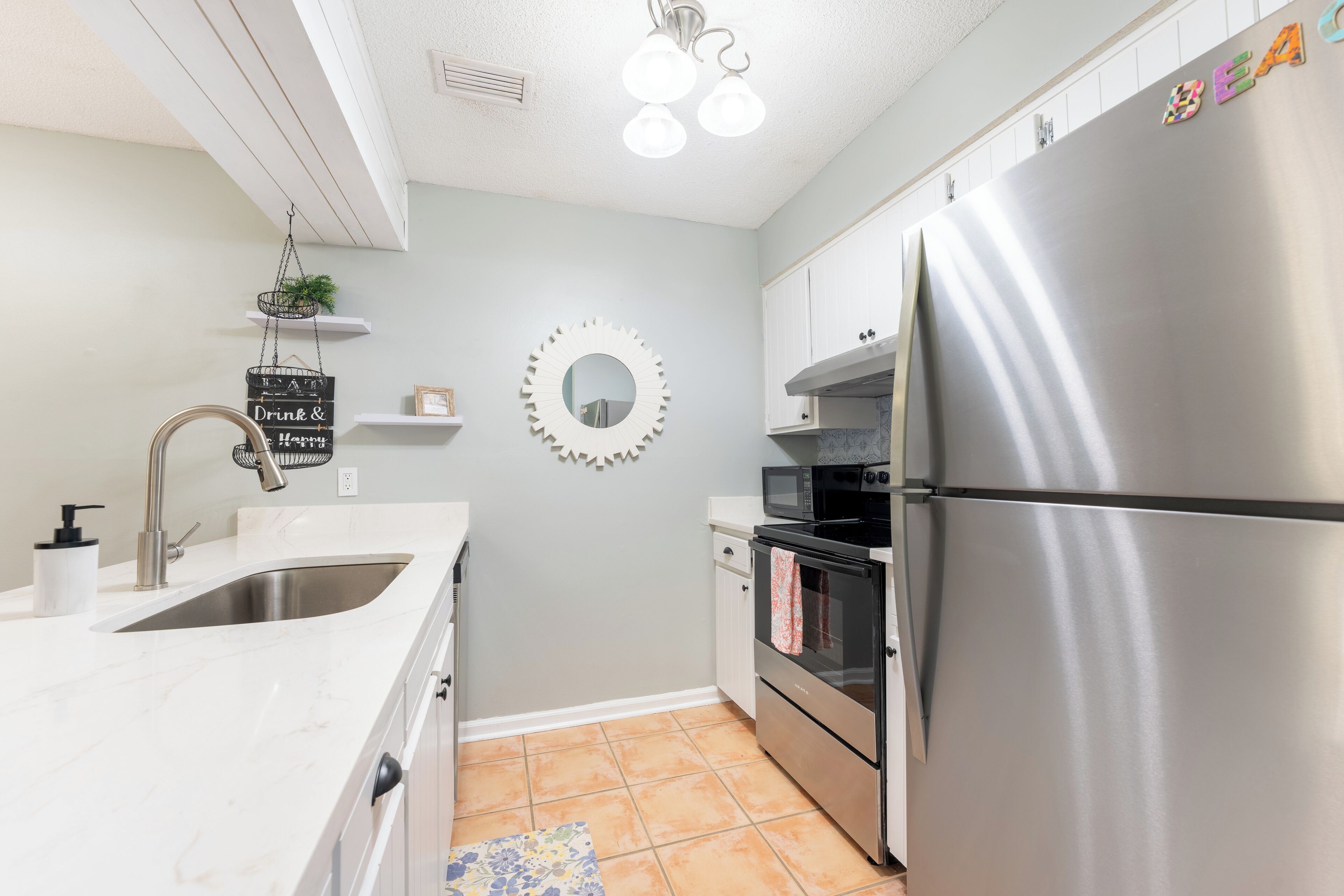 A modern kitchen with a stainless steel refrigerator, a built-in oven, and a microwave. There is a sink with a faucet, and a decorative mirror on the wall.