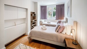 Een slaapkamer met twee bedden, een nachtkastje met een lamp, een raam met gordijnen, een inbouwkast en een plank met decoratieve spullen.
