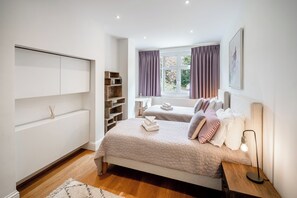 A bedroom with two beds, a bedside table with a lamp, a window with curtains, a built-in wardrobe, and a shelf with decorative items. - Family Serviced Apartment in Surrey (Claygate)