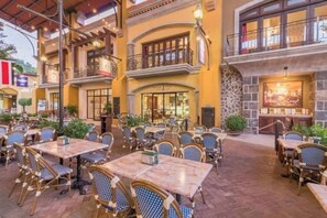 Ein Cafébereich im Freien mit mehreren Tischen und Stühlen, einem Steinkamin und einem Balkon mit Geländer.