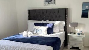 Una cama bien hecha con cabecera oscura y ropa de cama blanca, una mesita de noche con una lámpara, y un cuadro en la pared.