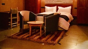 A bedroom with a bed, two chairs, a small table, a rug, and a cityscape painting on the wall. - Riad Ait Ben Haddou (Aït Benhaddou)