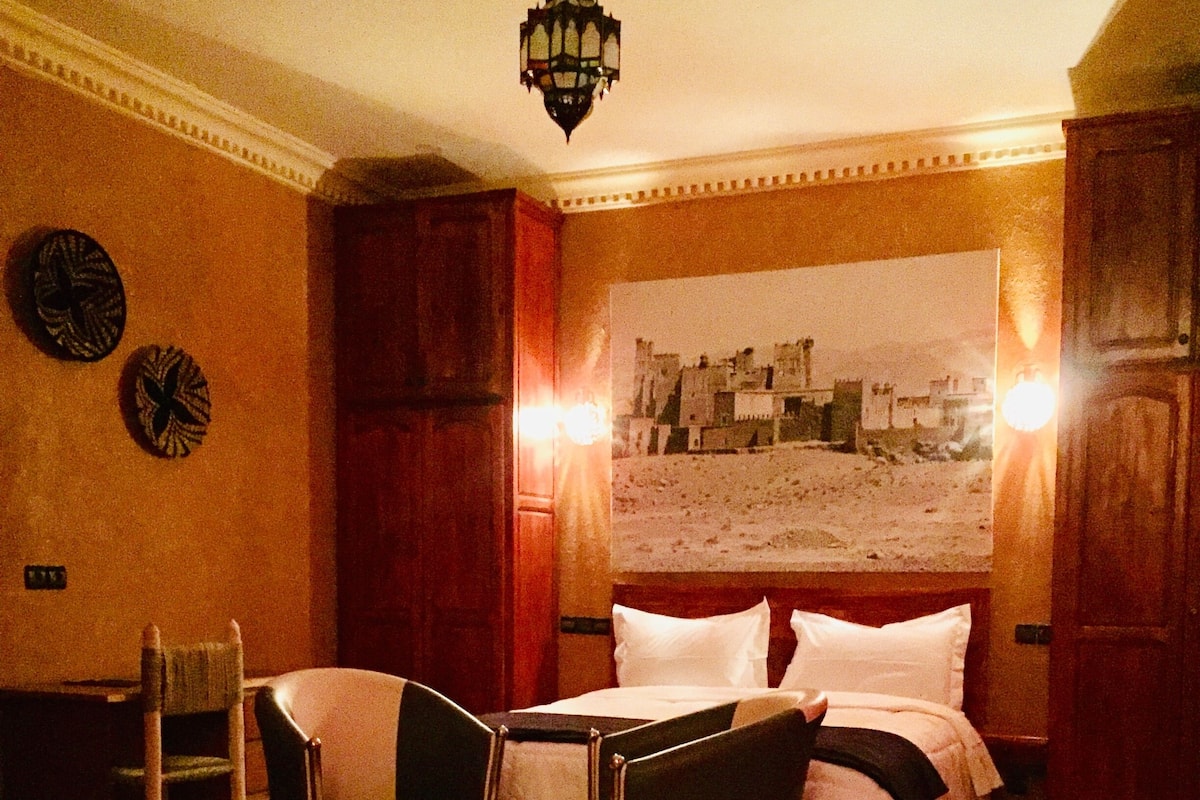 Quarto com cama, duas cadeiras, uma mesinha, tapete e pintura da paisagem urbana na parede.