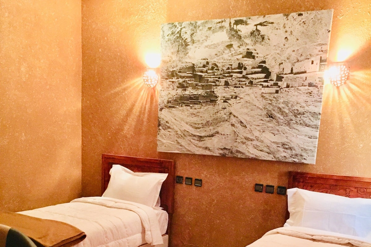 Quarto de hotel com duas camas de solteiro, cabeceira de madeira, uma mesa pequena e uma cadeira.