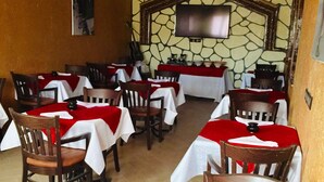 A restaurant with multiple tables covered in red tablecloths, wooden chairs, and a stone wall with a television. - Riad Ait Ben Haddou (Aït Benhaddou)