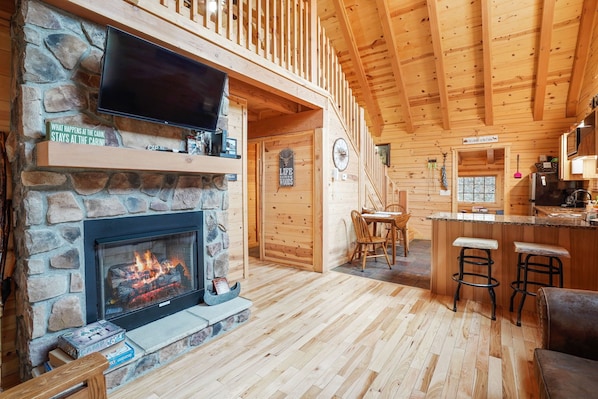 A cabin interior with a stone fireplace, wooden walls, a television mounted above it, and a bar area with stools.