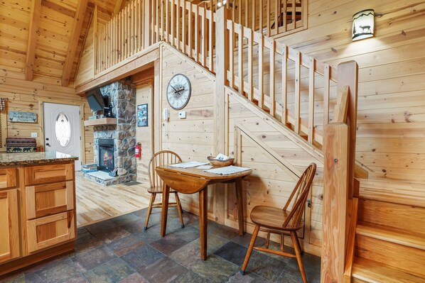 A wooden cabin interior with a stone fireplace, wooden dining table, and a staircase.