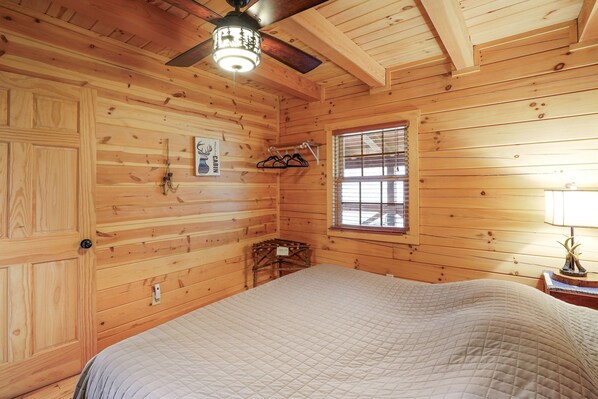Una habitación tipo cabaña de madera con una cama, un ventilador de techo, una ventana con persianas y una mesita de noche con lámpara.