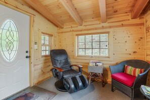 A wooden cabin interior with a massage chair, wicker chair with a red cushion, and a small table with books.
