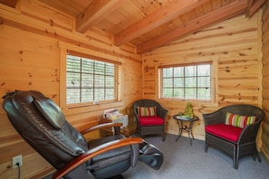 A log cabin interior with a leather chair, two wicker chairs with cushions, a small table, and a window with blinds.