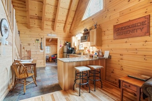 A wooden cabin interior with a kitchen area, a dining table with chairs, and a bar with stools. - Creekside Serenity | Hocking Hills, Creek, Hot Tub (Logan)