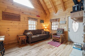 A cozy cabin interior with a wooden interior, a couch, a small table, a lamp, and a door with a window. - Creekside Serenity | Hocking Hills, Creek, Hot Tub (Logan)