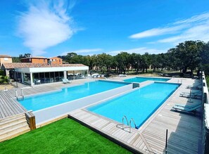 Une grande piscine extérieure dotée d’une terrasse en bois et de chaises longues, entourée d’arbres et bordée d’un bâtiment au toit recouvert de tuiles.