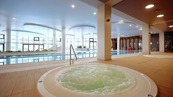 A large indoor swimming pool with a hot tub in the center, surrounded by a spacious area with high ceilings and large windows.