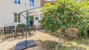 En udendørs terrasse med et bord og stole, et lille træ og en bygning i baggrunden.