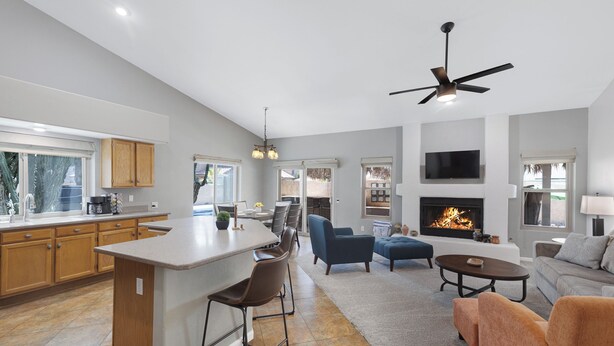 A modern kitchen with a central island, a dining area with chairs, a living room with sofas, a fireplace, and a ceiling fan. - Pool Home Near Downtown Gilbert! (Gilbert)