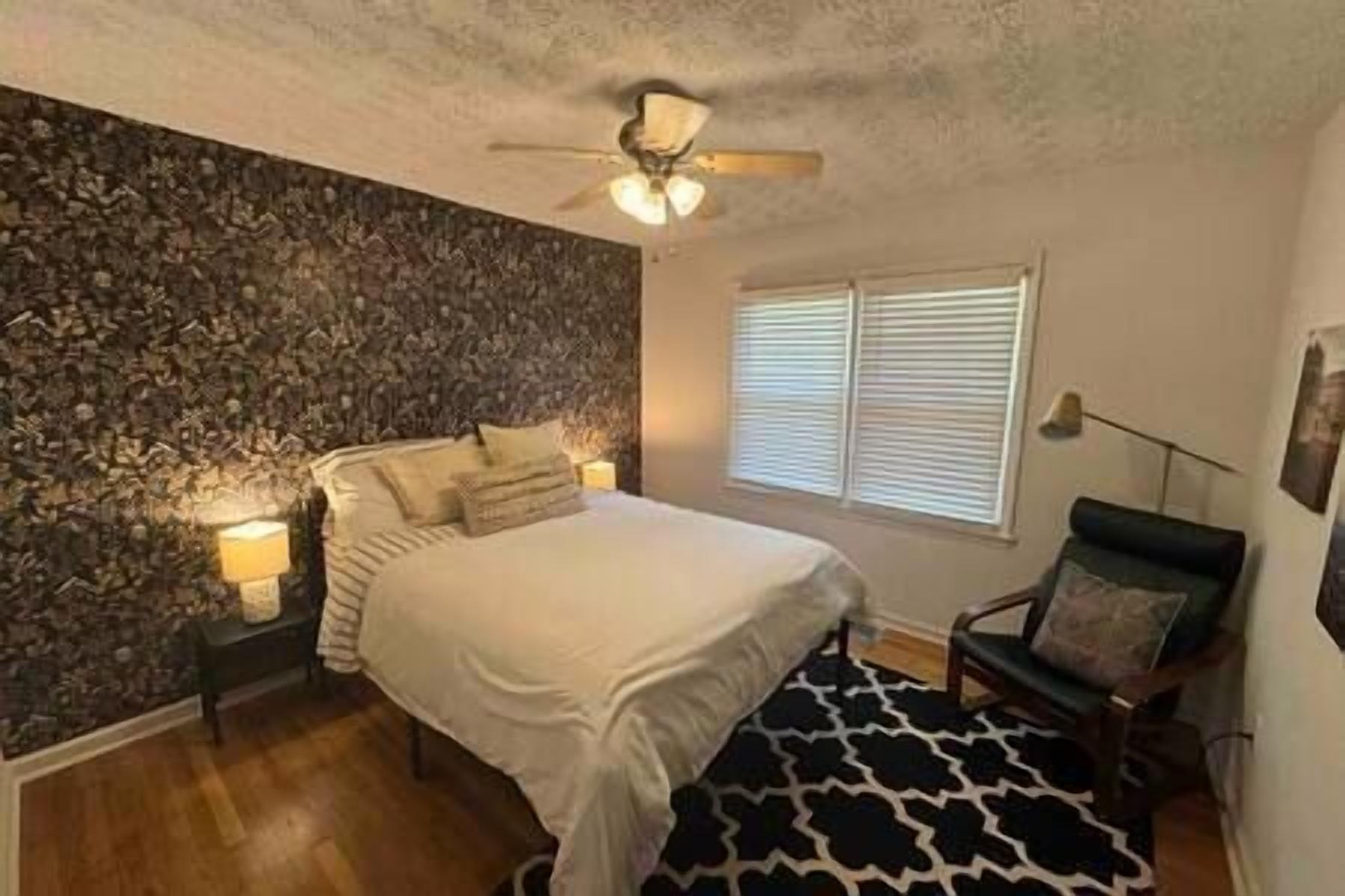 A bedroom with a stone wall, a bed with white bedding, a ceiling fan, and a window with blinds.