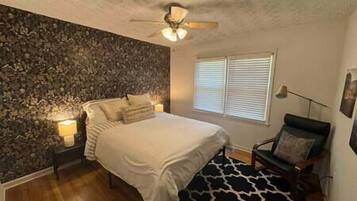 A bedroom with a stone wall, a bed with white bedding, a ceiling fan, and a window with blinds.