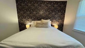 Een net opgemaakt bed met een hoofdbord, twee bedlampjes en een raam met jaloezieën.