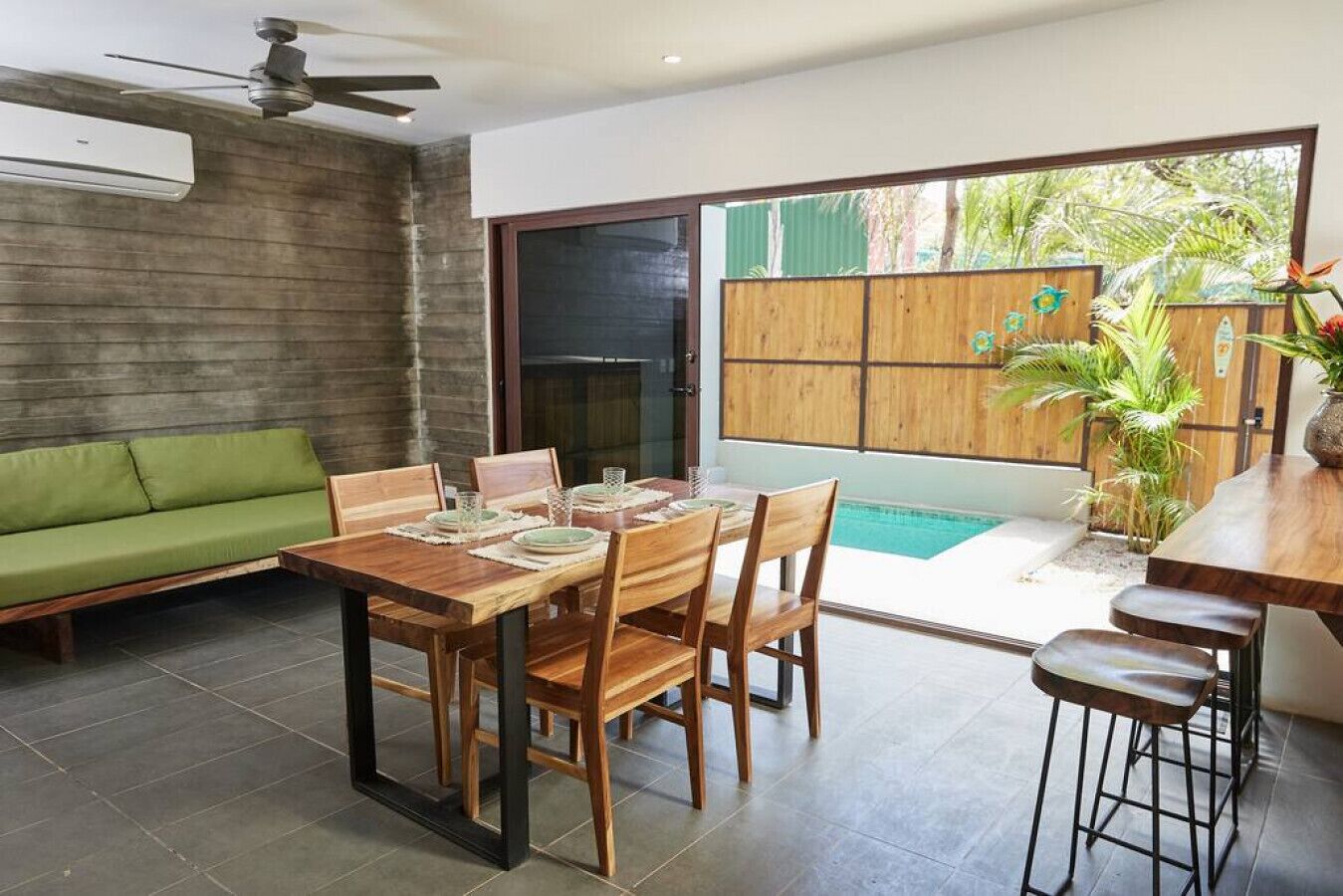 Un área de comedor con una mesa y sillas de madera, un sofá verde y vistas a una piscina por un tragaluz corredera.