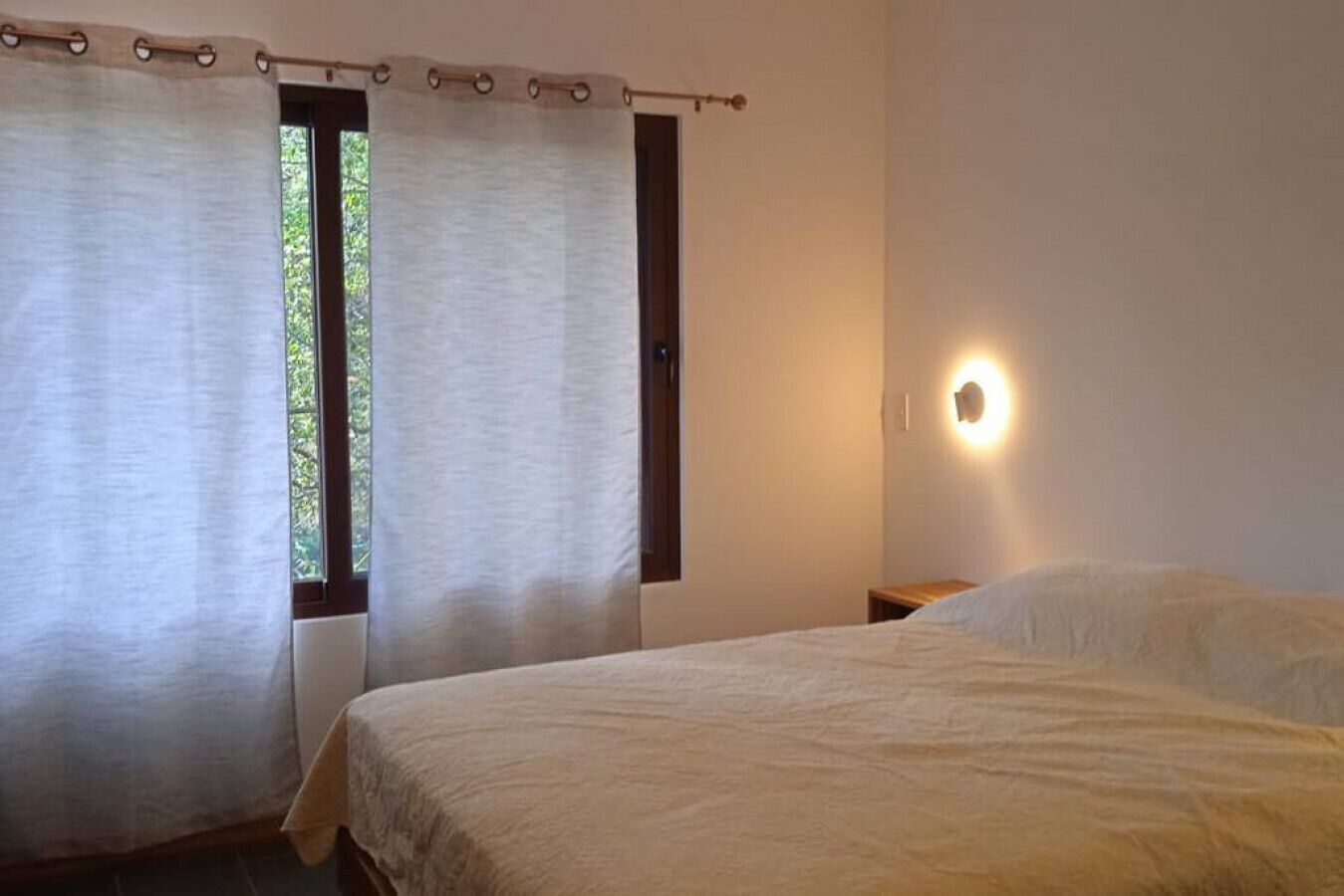 Ein Schlafzimmer mit einem Bett, einem Fenster mit Vorhängen, einem Nachttisch und einer Wandleuchte.