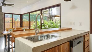 Una cocina moderna con lavavajillas integrado, un ventanal con vistas a una terraza de madera y follaje, y un ventilador de techo.