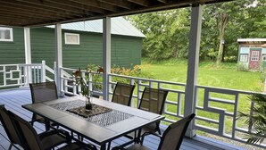 A covered outdoor patio with a table and chairs, a green house in the background, and a grassy yard.
