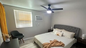 Une chambre à coucher comprenant un lit, des tables de chevet, un ventilateur de plafond, une fenêtre avec des stores, une chaise et un bureau.