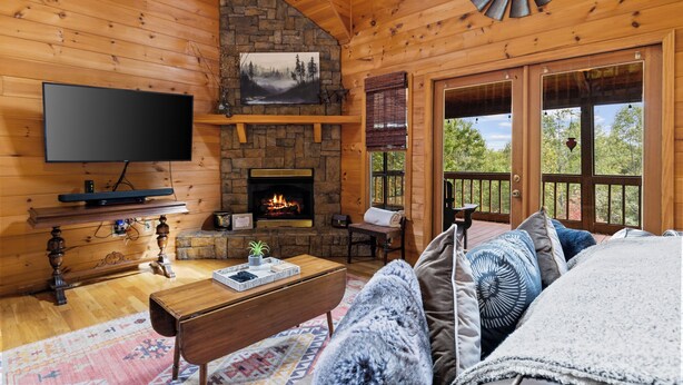 Ein gemütliches Blockhaus-Interieur mit Kamin aus Stein, einem Flachbildfernseher, einer bequemen Couch und einem großen Fenster mit Blick ins Freie.
