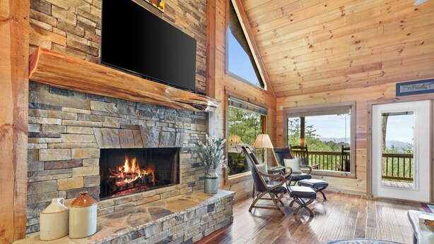 Une cheminée en pierre avec un feu de bois, un plancher en bois et une vue sur l’extérieur.