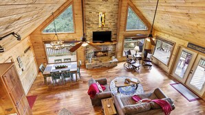 Un vaste espace de vie de style loft, doté d’une cheminée en pierre, de planchers en bois et de grandes fenêtres offrant une vue sur l’extérieur.