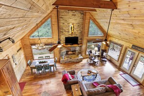 A spacious loft-style room with a stone fireplace, wooden floors, and large windows offering views of the outdoors.