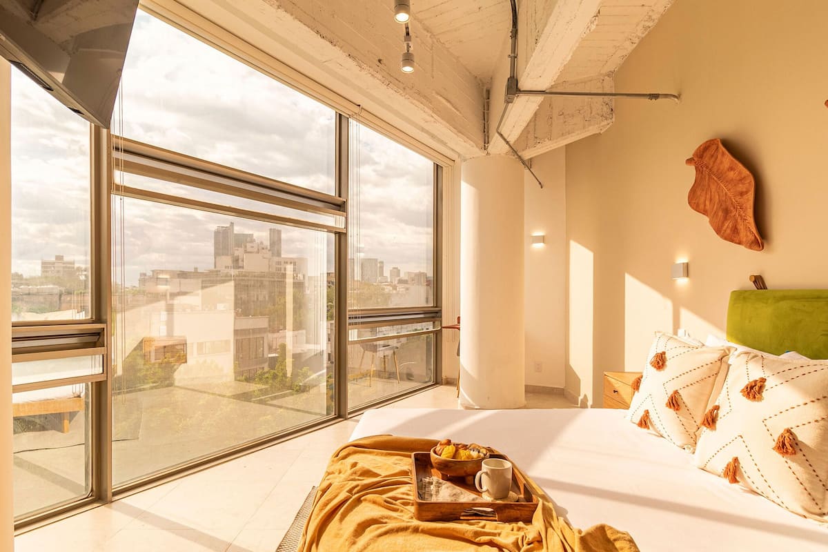 A modern hotel room with large windows, a city view, a green headboard, and a breakfast tray on the bed.