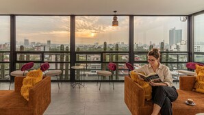A spacious room with large windows offering a city view, a comfortable seating area with chairs and a sofa, and a person reading a book. - Kukun Insurgentes Roma (Mexico City)