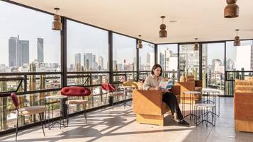 Un balcón espacioso con vista a la ciudad, amoblado con sillas y mesas modernas, y una persona leyendo un libro.