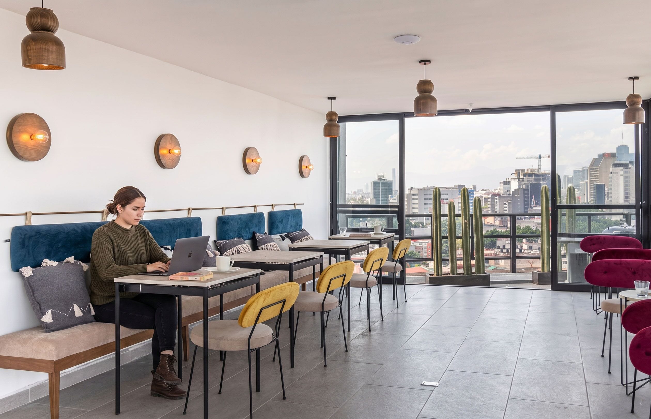 Un interior moderno con vistas a la ciudad, que incluye una zona de estar con mesas y sillas, una persona usando una computadora portátil, e iluminación colgante.