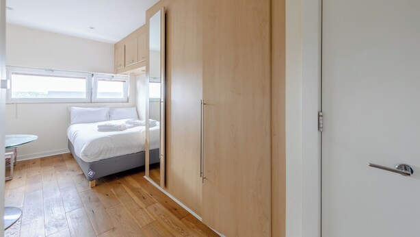 A bedroom with a bed, a wardrobe, a small table, and a window. - Modern 1BD Flat - 5 min walk to Battersea Park! (London)