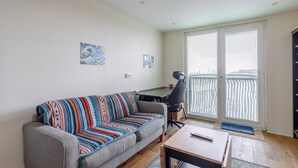 A living room with a gray sofa, a wooden coffee table, and a black office chair. There are two windows with blinds, a door, and a balcony with a railing. - Modern 1BD Flat - 5 min walk to Battersea Park! (London)