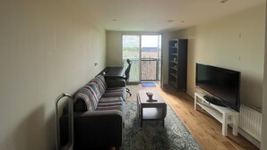 A living room with a sofa, a coffee table, a TV on a stand, and a balcony with a view. - Modern 1BD Flat - 5 min walk to Battersea Park! (London)