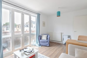 Ein modernes Wohnzimmer mit einer Couch, einem Sessel, einem Tisch mit Büchern und einer Fernbedienung sowie Blick durch große Fenster.
