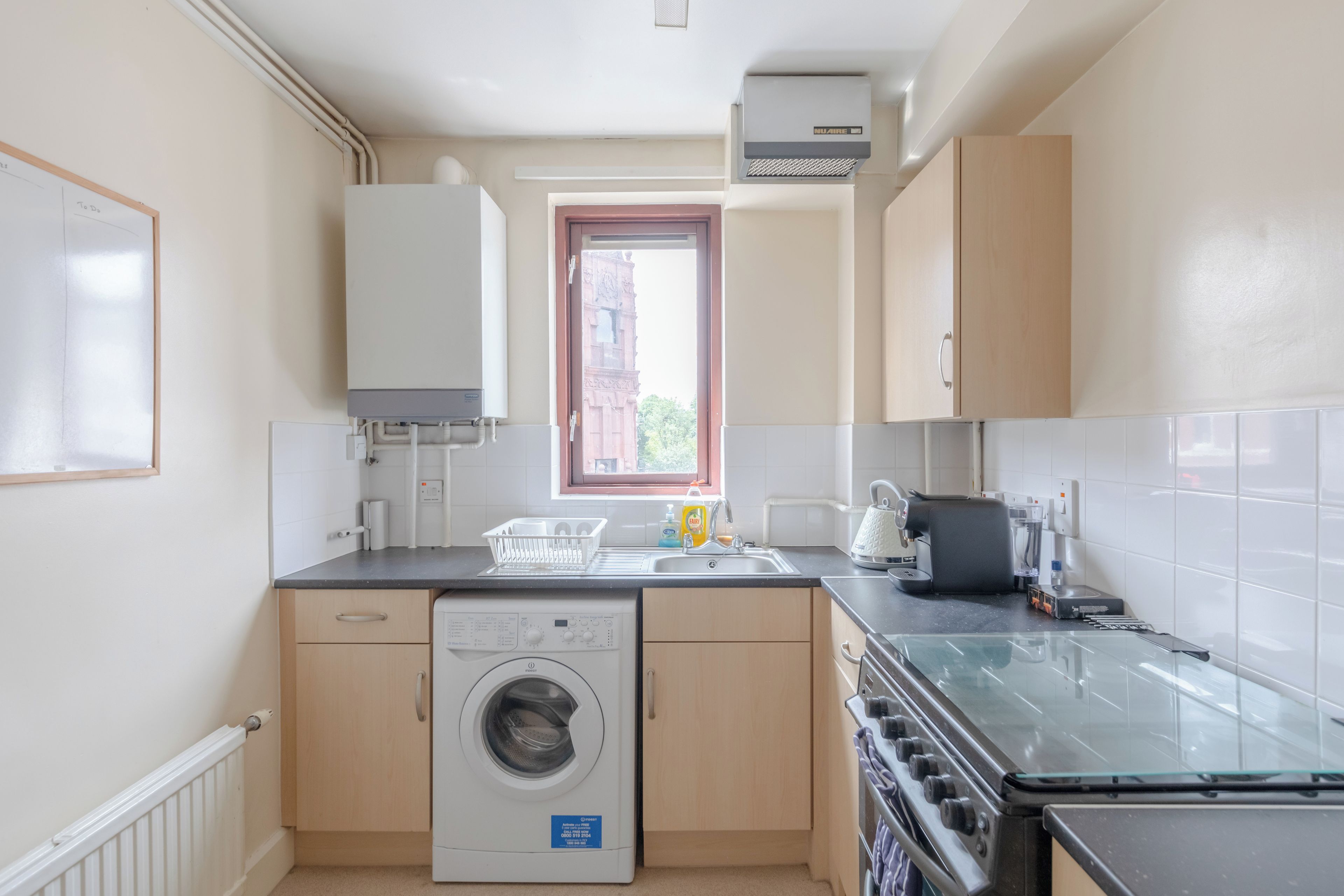 A compact kitchen with a washing machine, a coffee machine, and a window with a view of a building outside.