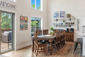 A spacious dining area with a wooden table and chairs, a kitchen with a coffee station, and artwork on the walls. - New Lakefront Cabin - Lake Views - Large Hot Tub - Firepit  (Branson)