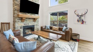 A living room with a stone fireplace, leather sofas, a wooden coffee table, and a large window overlooking a deck.