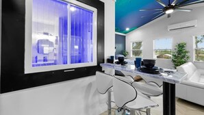 Une cuisine moderne avec une grande fenêtre, des tabourets de bar blancs, un ventilateur de plafond et un canapé blanc.
