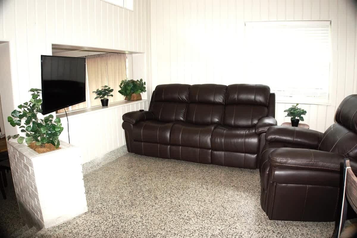 A living room with a brown leather sofa, a flat-screen TV mounted on the wall, and two potted plants.