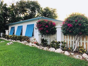 Una casa de un solo piso con fachada blanca y contraventanas azules, rodeada de césped cuidado y plantas con flores.