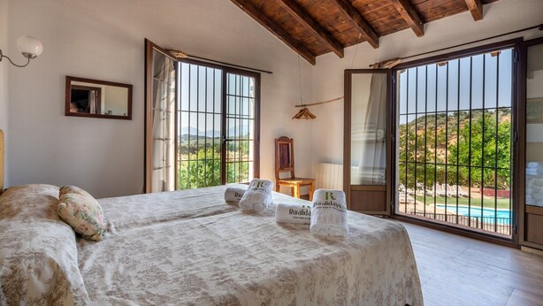 Ein Schlafzimmer mit einem Bett, zwei Handtüchern, einem Sessel, einer Hängelampe und Blick auf Pool und Landschaft.