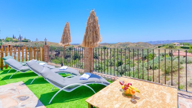 Un balcon avec deux fauteuils, une table garnie de fruits et une vue sur un paysage vallonné.