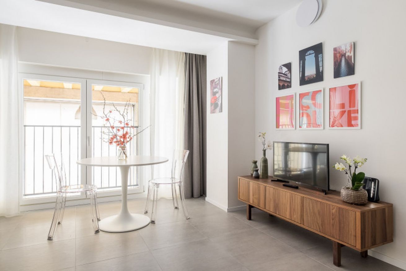 A modern living room with a round dining table, clear chairs, a wooden TV stand, and artwork on the wall.