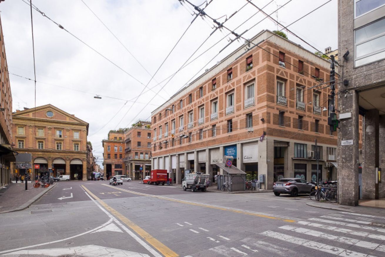 A street view with a building featuring a large arched entrance, a red van parked on the street, and a tram line overhead.